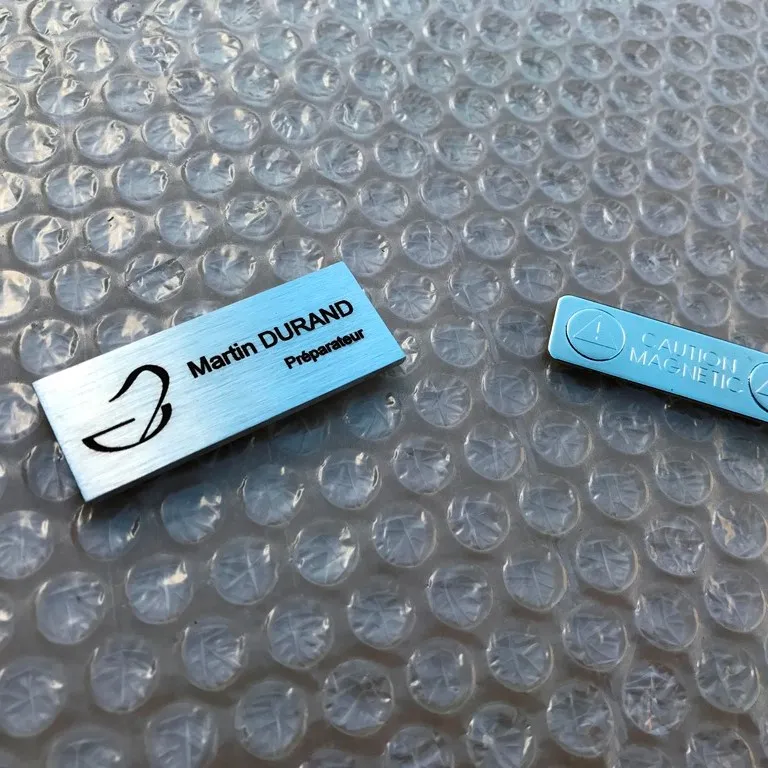Badge nominatif en inox brossé gravé au laser avec logo, présenté avec son attache magnétique séparée, posé sur film à bulles