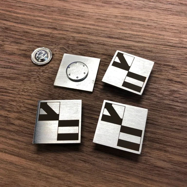 Badges carrés en inox brossé gravés au laser d'un logo géométrique, l'un retourné montrant sa fixation magnétique et son aimant séparé, posés sur un plan en bois foncé