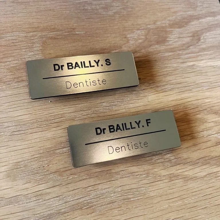 Deux badges nominatifs effet laiton brossé pour dentistes, gravés "Dr Bailly" avec la mention Dentiste