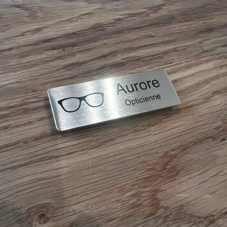 Badge nominatif en inox brossé gravé au laser pour opticienne, avec pictogramme de lunettes et la mention Aurore Opticienne
