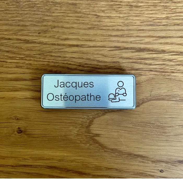 Badge nominatif en plastique effet aluminium brossé avec pictogramme d'ostéopathe et bordure grise, posé sur un plan en bois clair