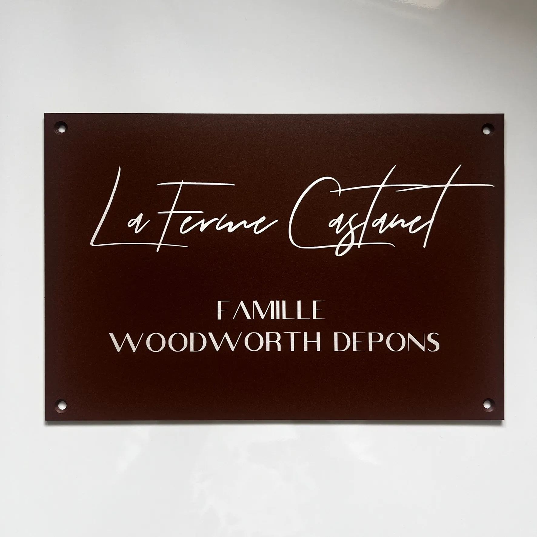 Plaque nominative rectangulaire de couleur brune avec texte blanc "La Ferme Castanet - Famille Woodworth Depons", percée aux quatre coins pour fixation murale