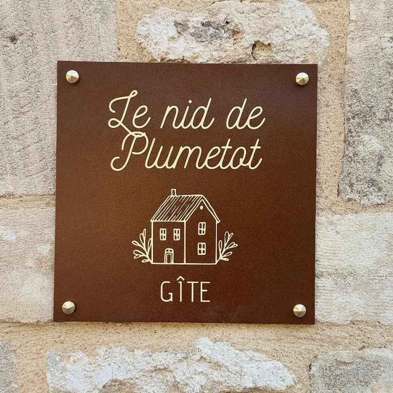 Plaque carrée de gîte en finition thermolaquée effet rouille, gravée d'un logo maison et d'un texte doré, fixée sur un mur en pierre par des vis à tête laiton