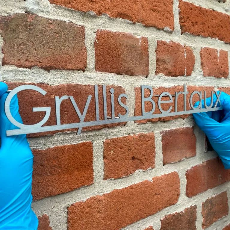 Enseigne de maison « Gryllis Bertaux » en lettres découpées inox tenue devant un mur de briques rouges