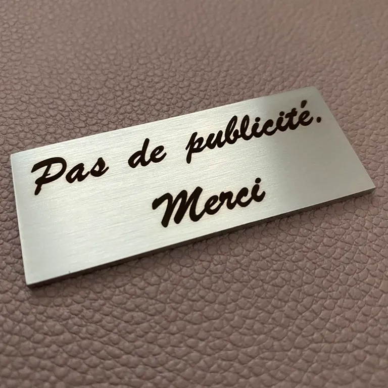 Petite plaque en inox brossé gravée "Pas de publicité, merci" en écriture manuscrite, posée sur un fond en cuir