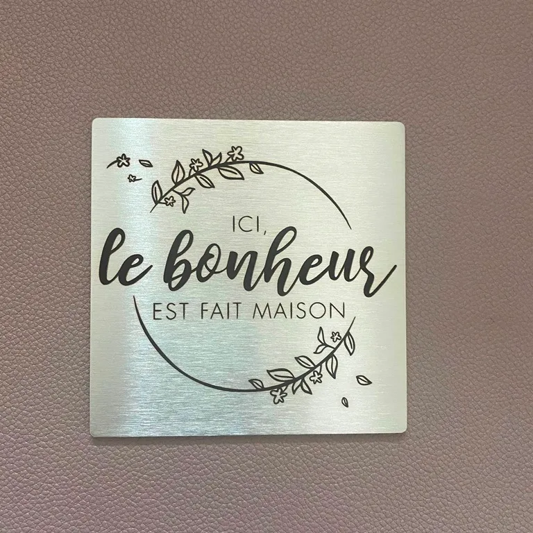 Plaque décorative carrée en inox brossé gravée au laser d'une citation "Ici, le bonheur est fait maison" entourée d'une couronne florale, posée sur un fond en cuir