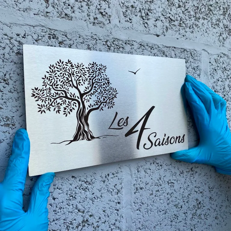 Plaque en inox brossé gravée au laser d'un olivier et du nom "Les 4 Saisons", tenue par des mains gantées contre un mur en pierre