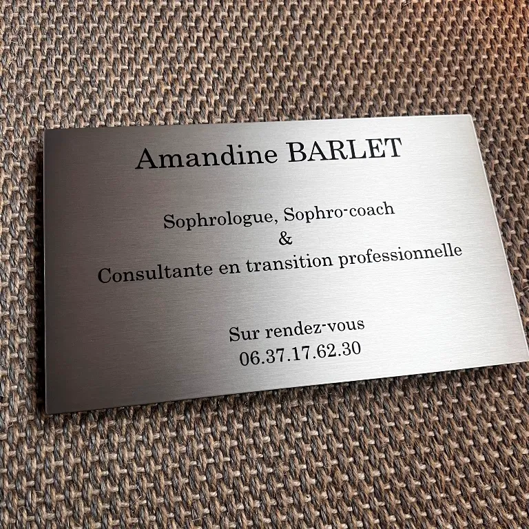 Plaque professionnelle en inox brossé gravée en noir pour une sophrologue et consultante, avec nom, activité et téléphone, posée sur un tissu tressé