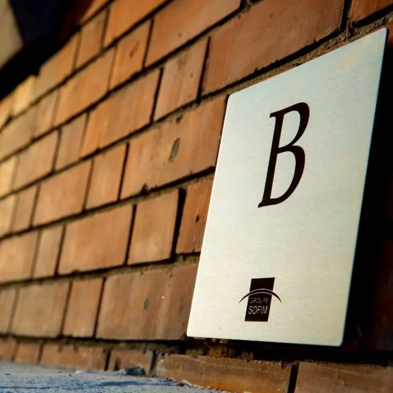Plaque signalétique en inox brossé gravée d'une lettre B et d'un logo d'entreprise, posée contre un mur en briques