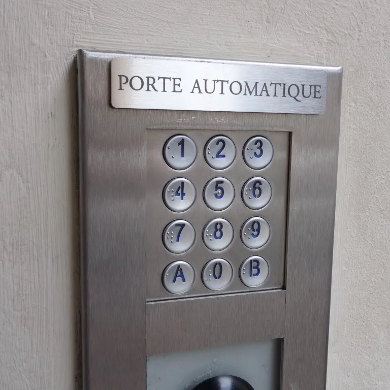 Plaque signalétique en inox brossé gravée "Porte automatique", fixée au-dessus d'un clavier à code sur un mur clair