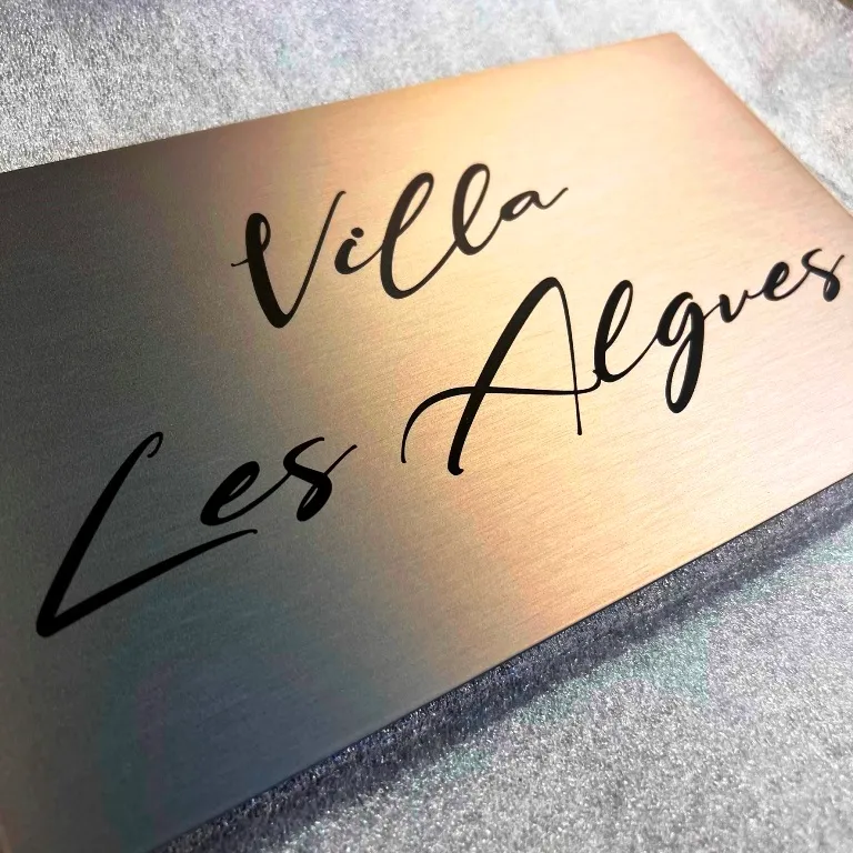 Plaque de maison en laiton brossé gravée au laser du nom d'une villa en écriture manuscrite, posée sur un tissu clair