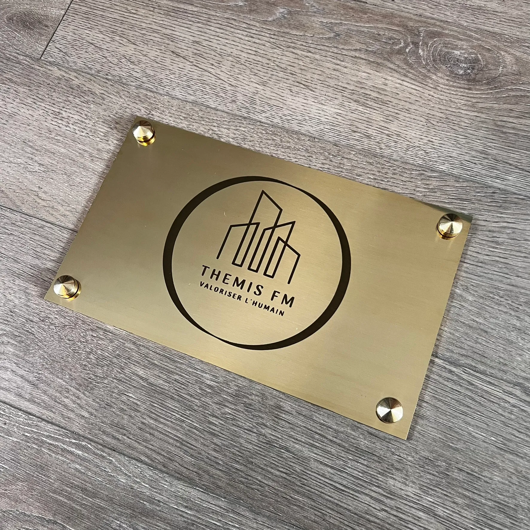 Plaque professionnelle en laiton brossé gravée au laser du logo et du slogan d'une entreprise, fixée par quatre entretoises laiton, posée sur un sol en bois