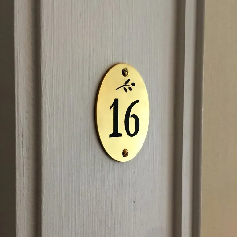 Plaque de porte ovale en laiton poli gravée du numéro 16 et d'un petit motif floral, vissée sur une porte claire