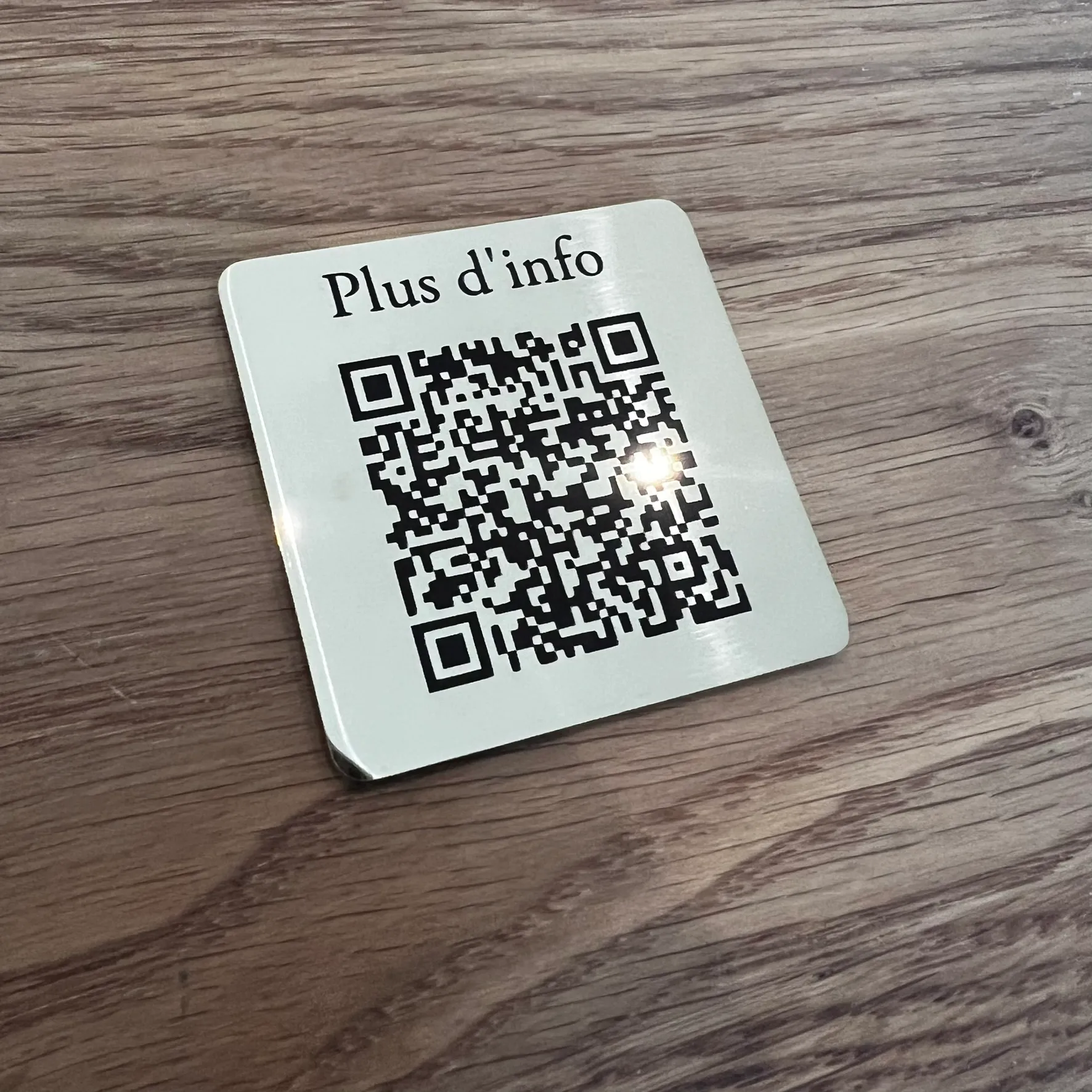 Plaque carrée en inox brossé posée sur bois, gravée au laser d'un QR code noir surmonté du texte « Plus d'info »