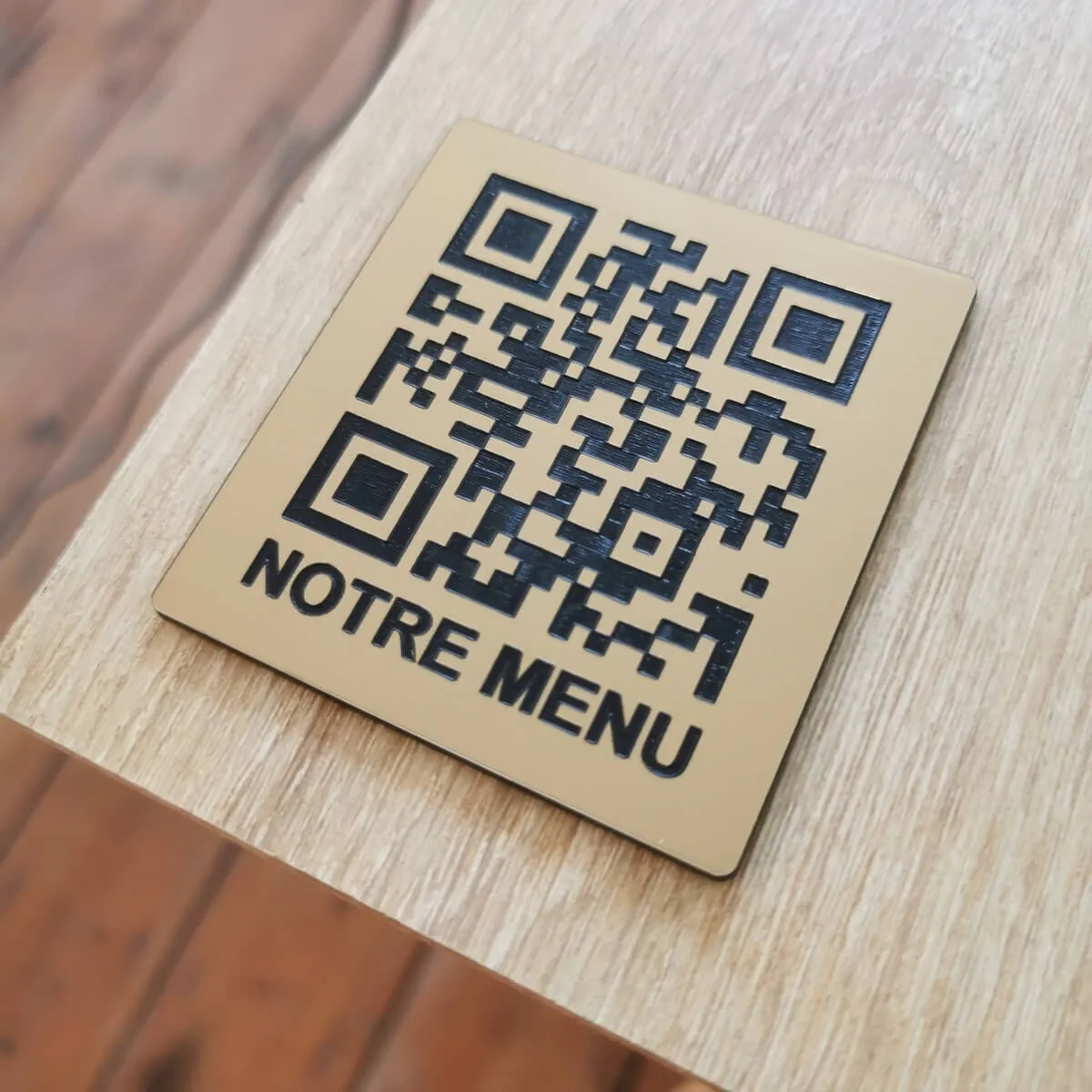 Plaque carrée en plastique effet laiton brossé posée sur bois, gravée au laser d'un QR code noir surmontant le texte « Notre menu »