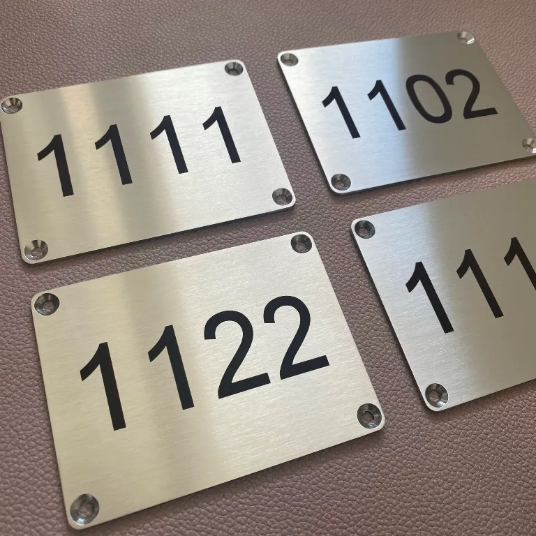 Plaques de numérotation en inox brossé gravées au laser de numéros noirs, percées aux quatre coins, posées sur un fond en cuir