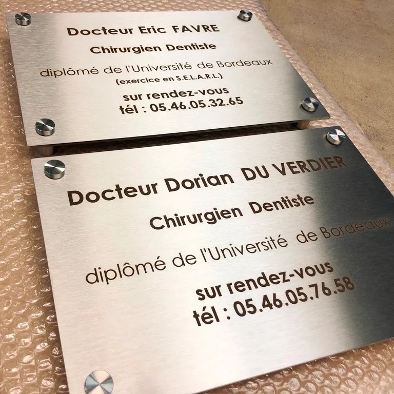 Deux plaques professionnelles en inox brossé gravées en noir pour des chirurgiens-dentistes, avec nom, diplôme et téléphone, fixées par des entretoises inox, posées sur film à bulles