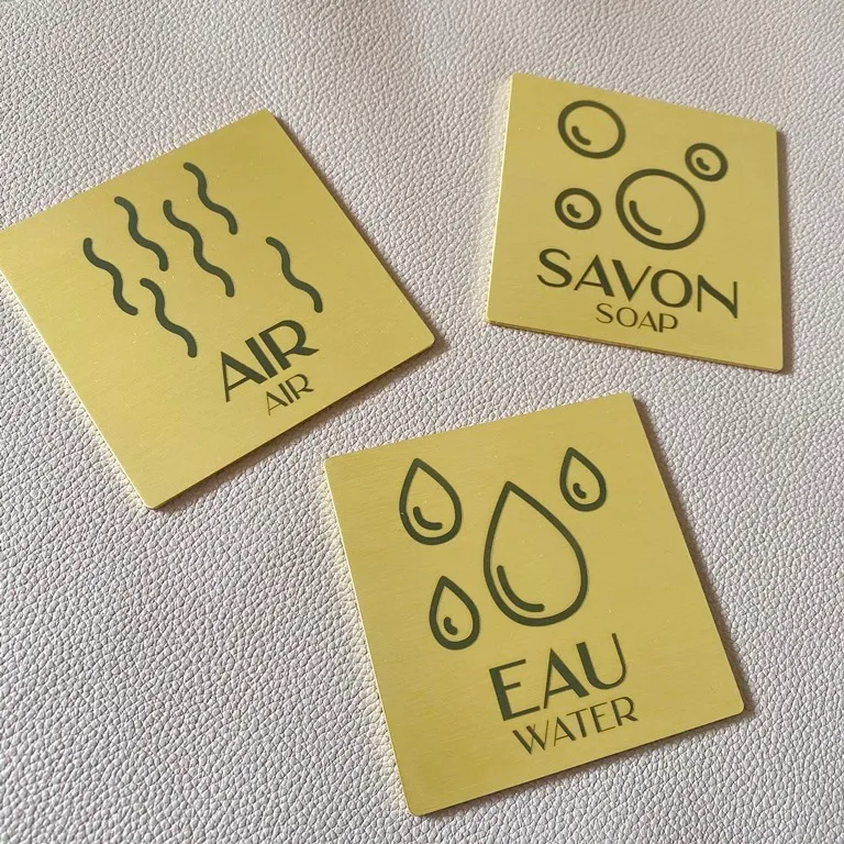Trois plaques carrées en laiton brossé gravées de pictogrammes et des mentions bilingues "Air", "Savon" et "Eau", posées sur un fond clair