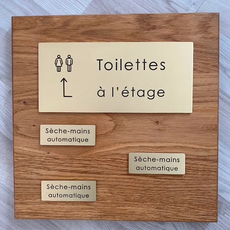 Plaques de signalétique en laiton brossé gravées "Toilettes à l'étage" avec pictogramme et flèche, et plusieurs plaques "Sèche-mains automatique", présentées sur un panneau en bois