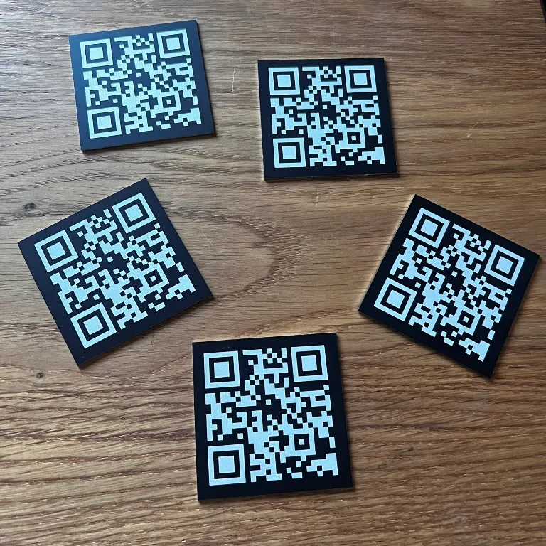 Cinq plaques carrées noir mat disposées sur bois, chacune gravée au laser d'un QR code dont le motif ressort en clair